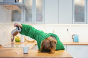 Exhausted woman with her head on kitchen counter pouring coffee and missing the cup.